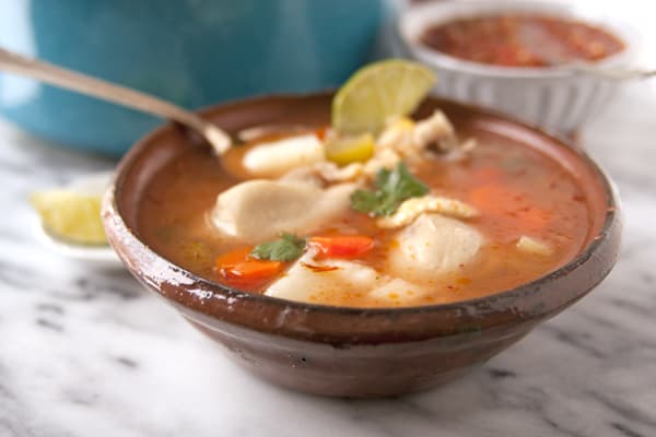 close up shot of caldo de pollow in a brown bowl with a silver spoon and slice of lime on a white marble table