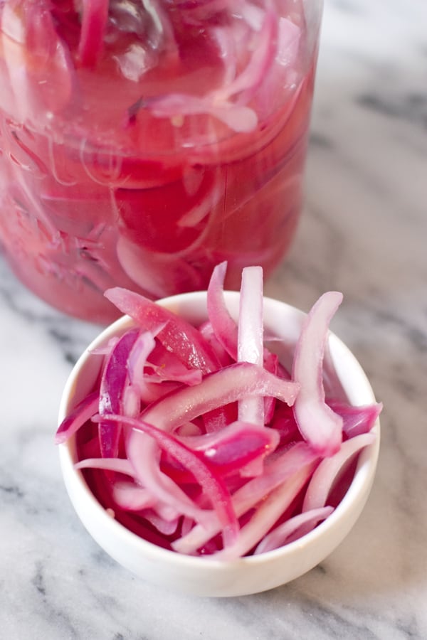Pickled Red Onions (a.k.a. Cebollas en Escabeche) in a small white ramekin with a mason jar filled with mexican pickled onions in the background.