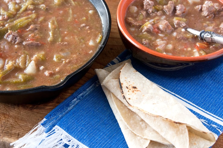 Chile Verde con Carne y Papas (Green Chile with Beef and Potatoes) in an iron skillet with a serving in a bowl and two rolled corn tortillas on a blue cloth napkin 