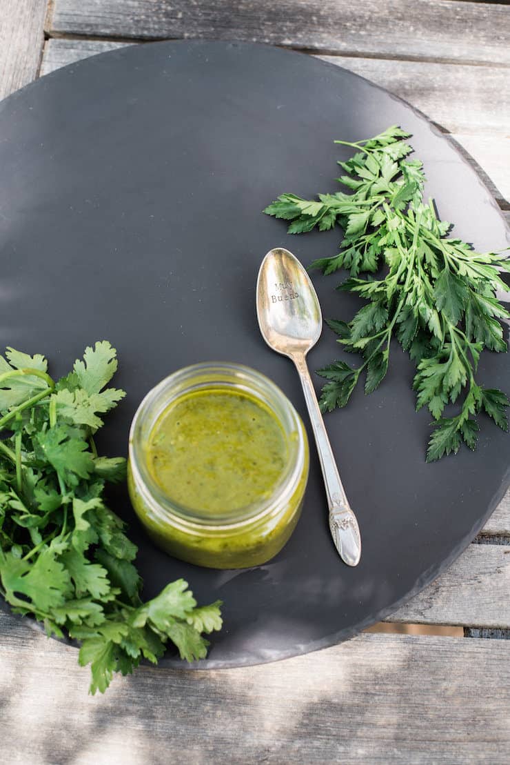 chimichurri sauce in a mason jar on a black plate with a spoon and cilantro and parsley on the side 