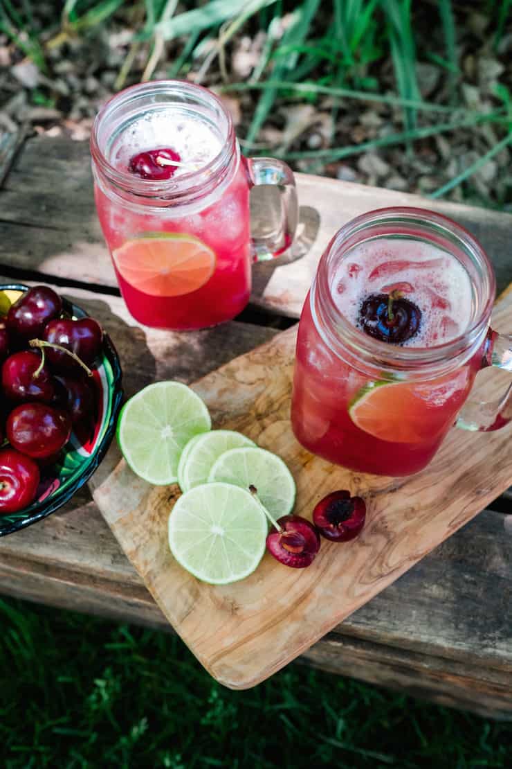 Mason jar mugs filled with Cherry Limeade Margaritas on a cutting board with line slices and bowl of cherries