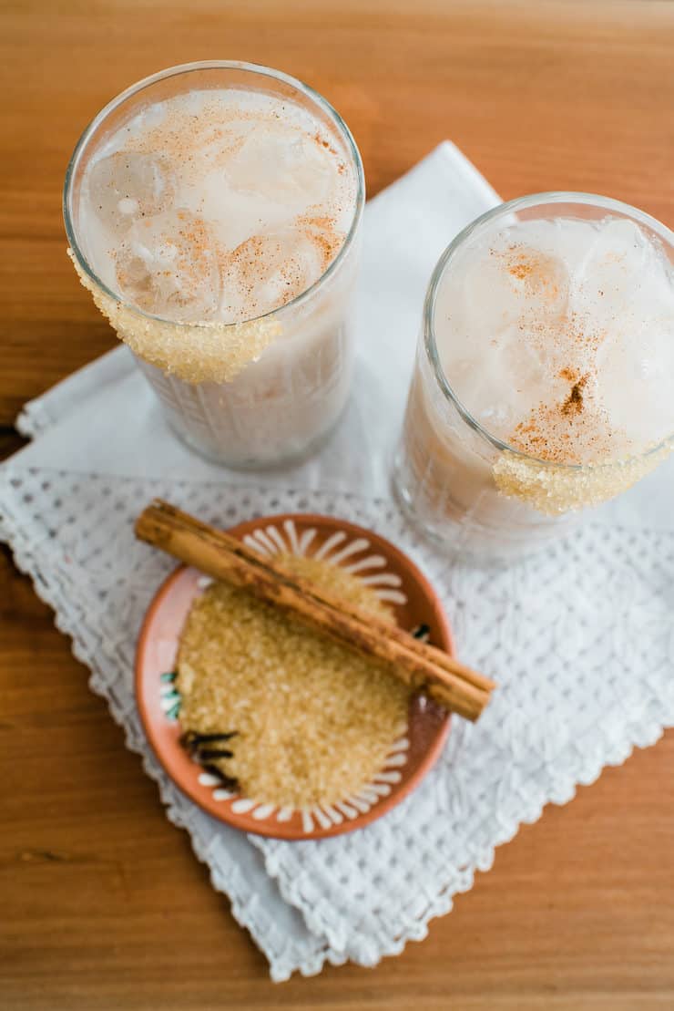 Two glasses filled with horchata margarita and a side bowl of raw sugar and a cinnamon stick