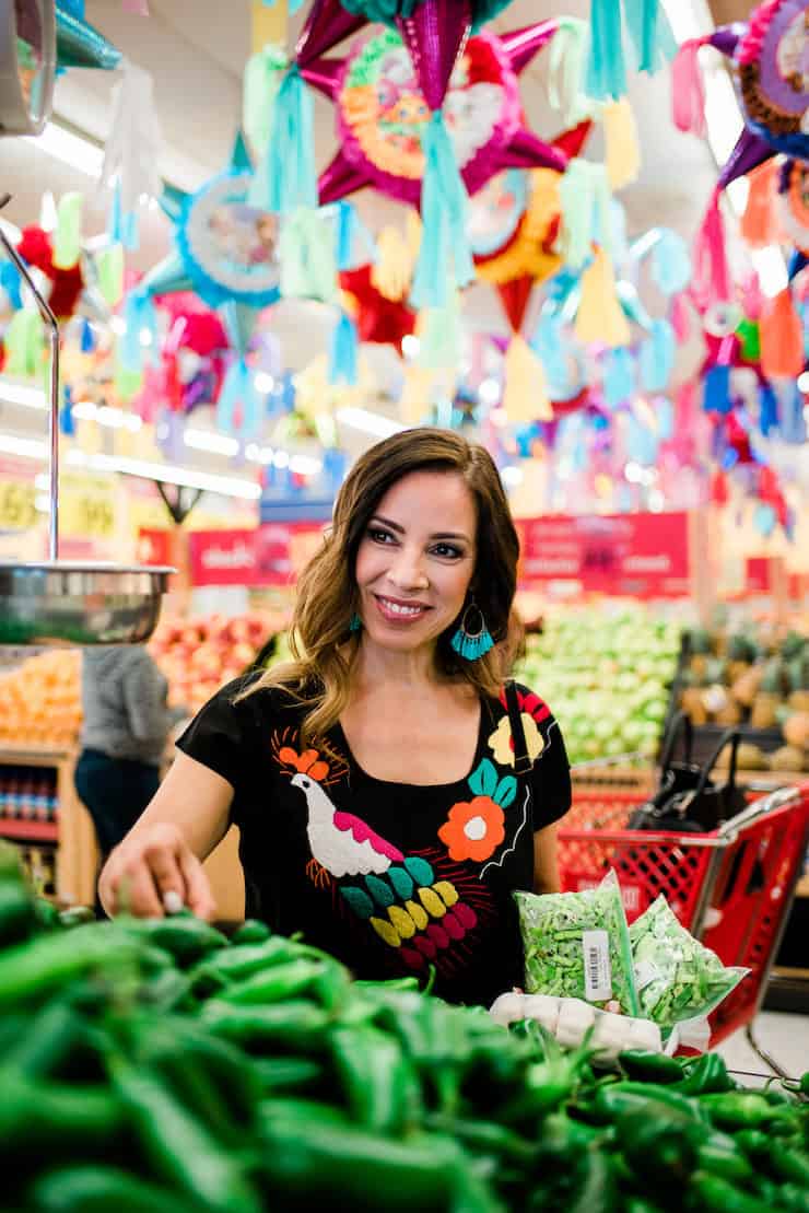 Mexican latina food blogger smiling and choosing jalapeños under ceiling lined with piñatas
