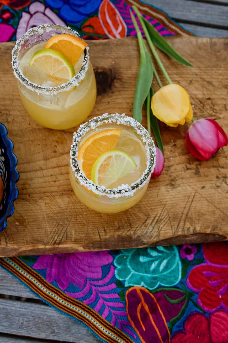 two skinny margaritas on a wooden cutting board with a colorful Mexican embroidered runner and yellow and pink tulips