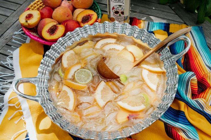 a pewter silver Mexican punch bowl filled with Paloma peach punch on a serape and basket of peaches and tequila in the background 
