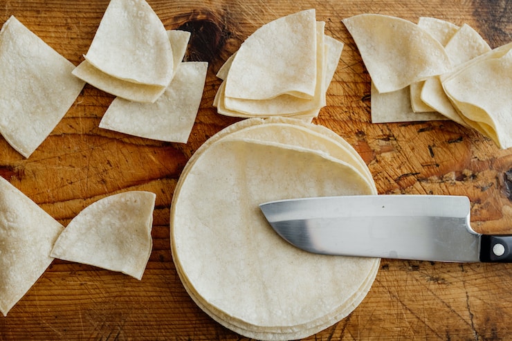 corn tortillas on a wooden cutting board with a chef's knife with some of the tortillas cut into quarters for frying 