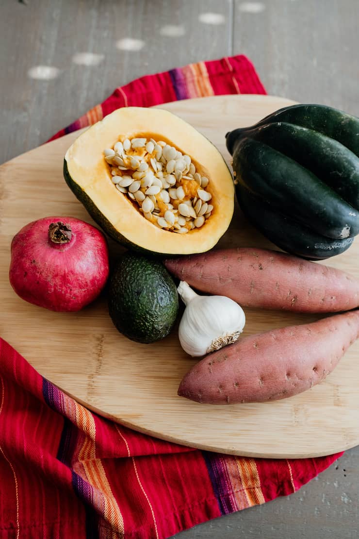 pomegranate, avocado, garlic, sweet potato, whole acorn squash and half of an acorn squash on a wooden cutting board on a red striped towel