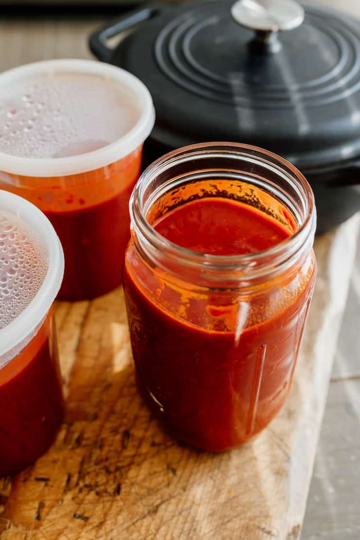 homemade Red Chile sauce in mason jar and freezer-safe plastic containers on a wooden board with a cast iron pot in the background 