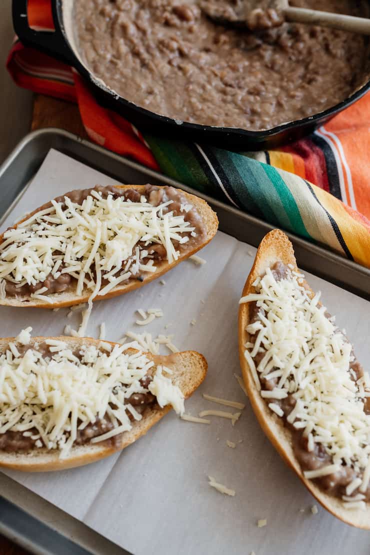 classic molletes on a parchment lined sheet pan prior to baking - bolillos with refried beans and shredded cheese