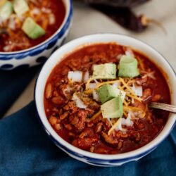 Mexican chili con carne topped with onions, cheese and avocados in blue and white bowls.