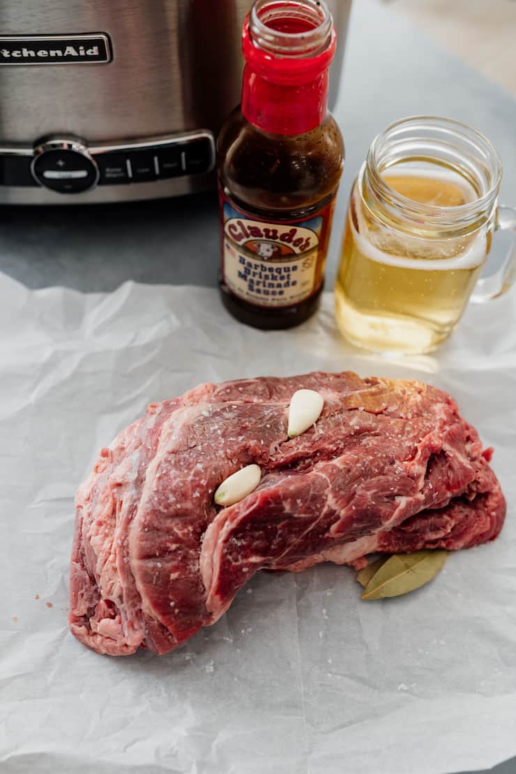 raw beef brisket on a piece of parchment in front of a slow cooker with a jar of claude's marinade and a glass of beer