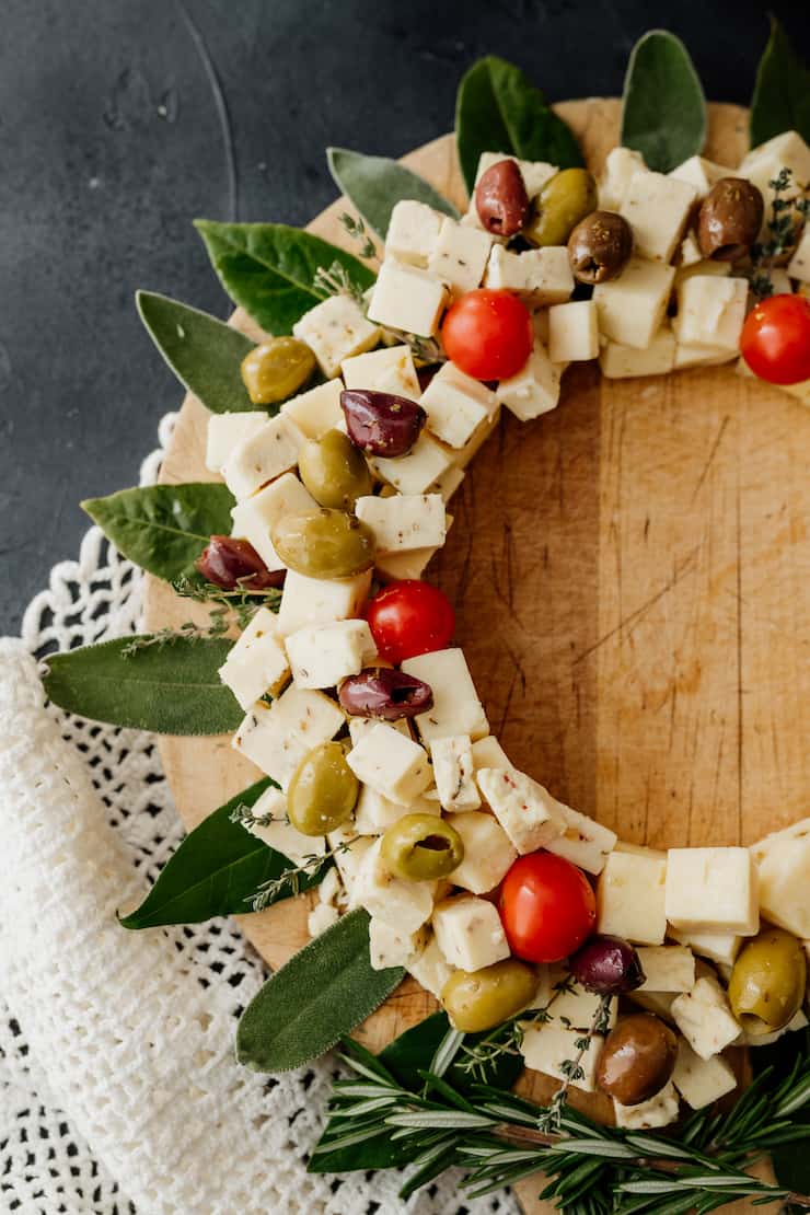 cheese cubes arranged in a wreath pattern with olives and tomatoes as ornaments and bay leaves as extra decor on a wooden cheese board