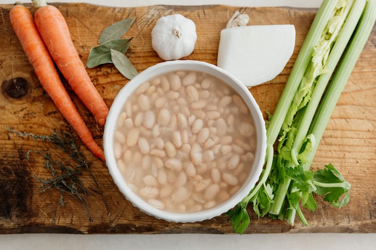 2 carrots, 3 bay leaves, a head of garlic, celery stalks and a bowl of white beans on a cutting board