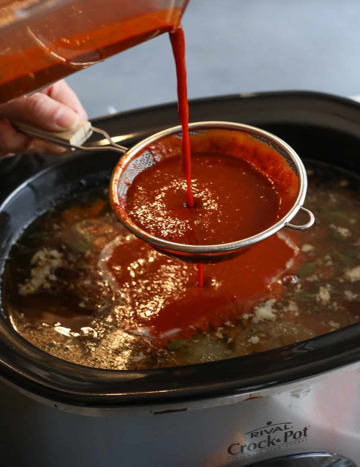 hand pouring homemade red chile sauce through a strainer into the crockpot with the birria