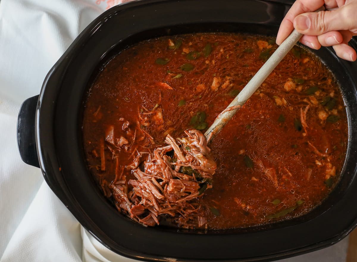 overhead shot peering down into bowl of slowcooker with a wooden spoonful of birria de res being pulled to the top
