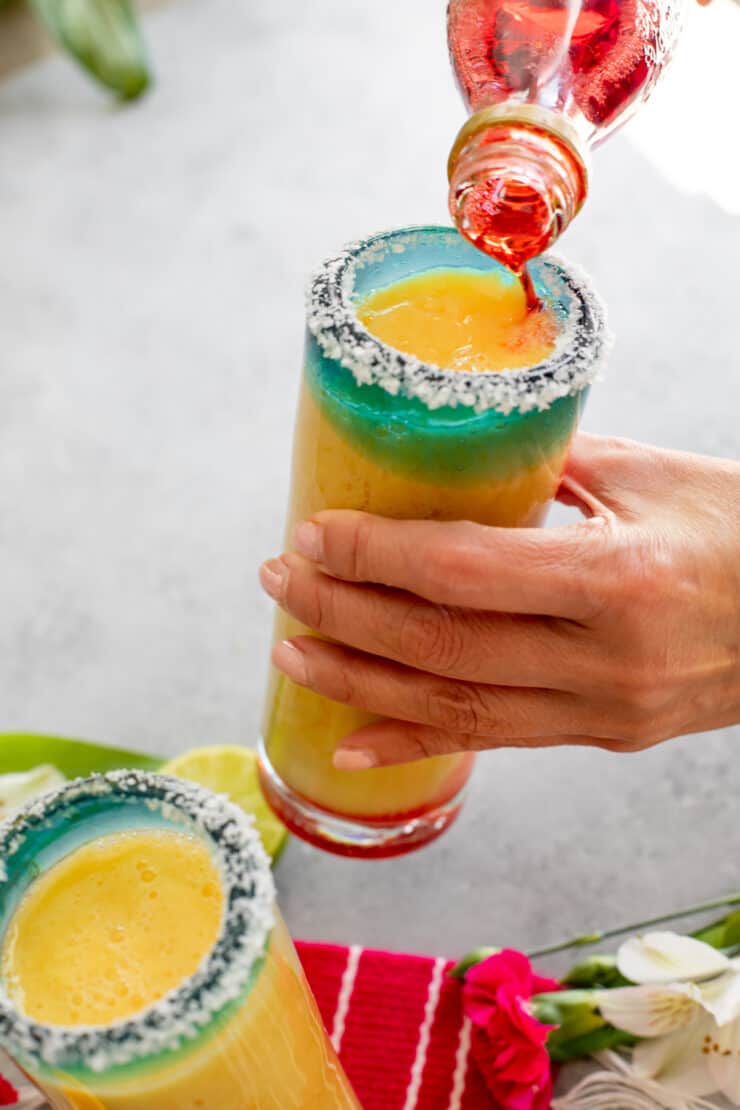 hand holding a collins glass filled with frozen orange margarita and pouring in the grenadine