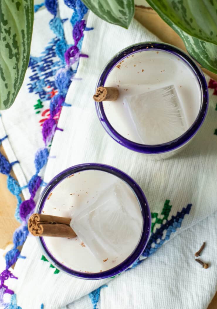 overhead shot of two glasses of horchata with cinnamon sticks on top of. an embroidered napkin