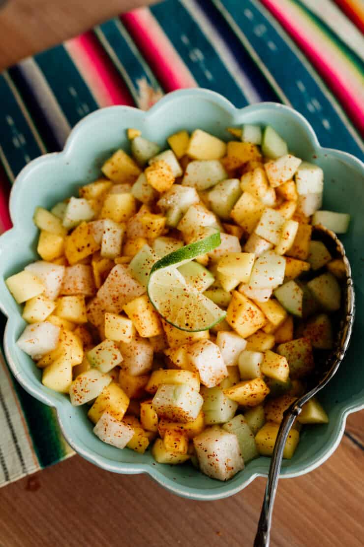 chopped mango jicama cucumber sprinkled with Tajin in a teal bowl on a Mexican textile runner