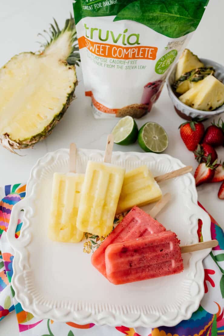 three pineapple paletas and two strawberry popsicles on a vintage platter and colorful Otomi fabric with fresh fruit around and a package of Truvia