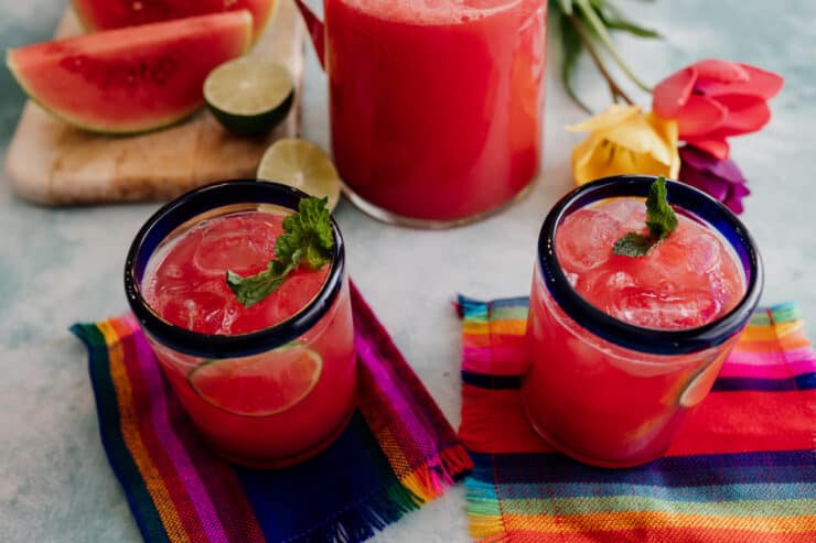 two glasses of agua de sandia with a pitcher of watermelon agua fresca in the background