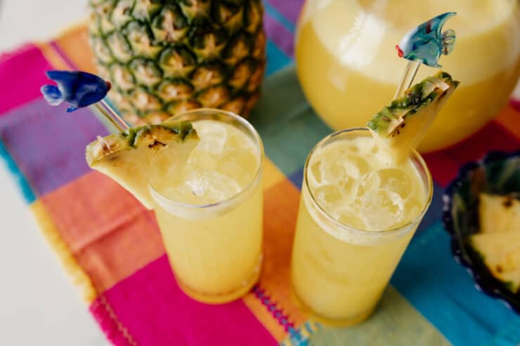 two collins glasses of pineapple agua fresca garnished with cocktail stirrers and fresh pineapple wedges