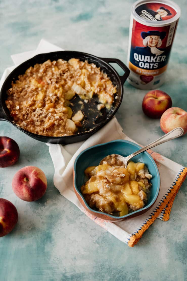 serving of peach crisp in a bowl and cast iron skillet with Quaker Oats and peaches in the background