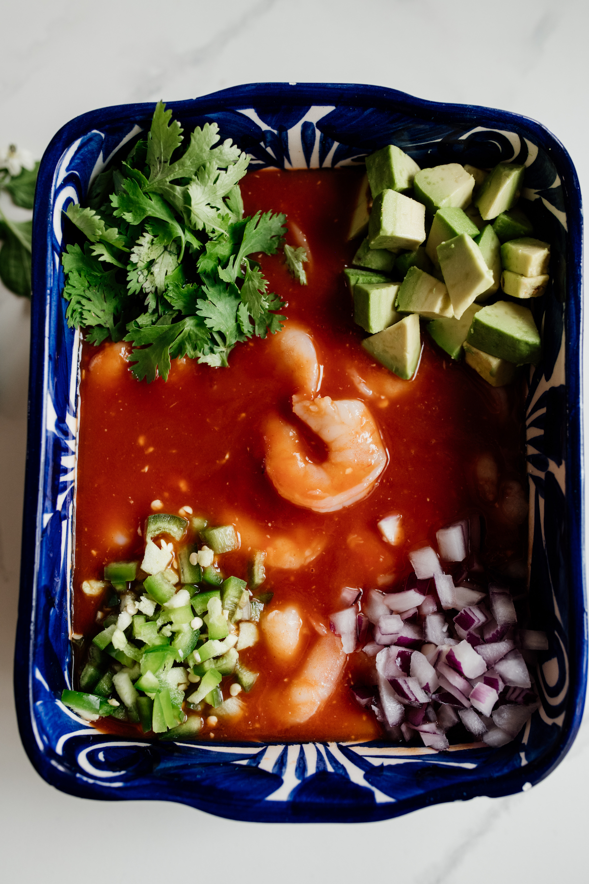 blue square bowl with shrimp in tomato sauce with corners of cilantro, avocado chunks, red onion and chiles that haven't been stirred in yet