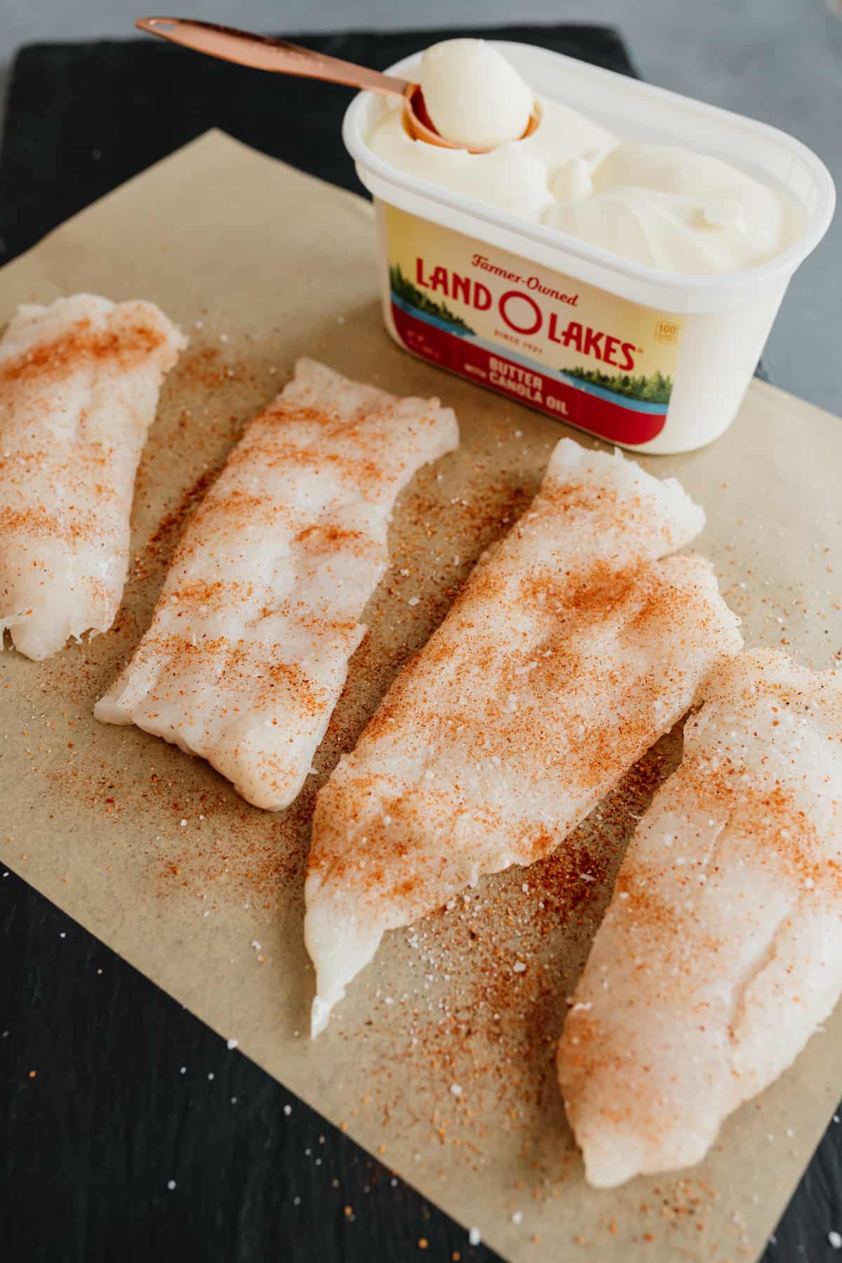 a tub of land o lakes butter and four cod fillets sprinkled with spice on parchment paper