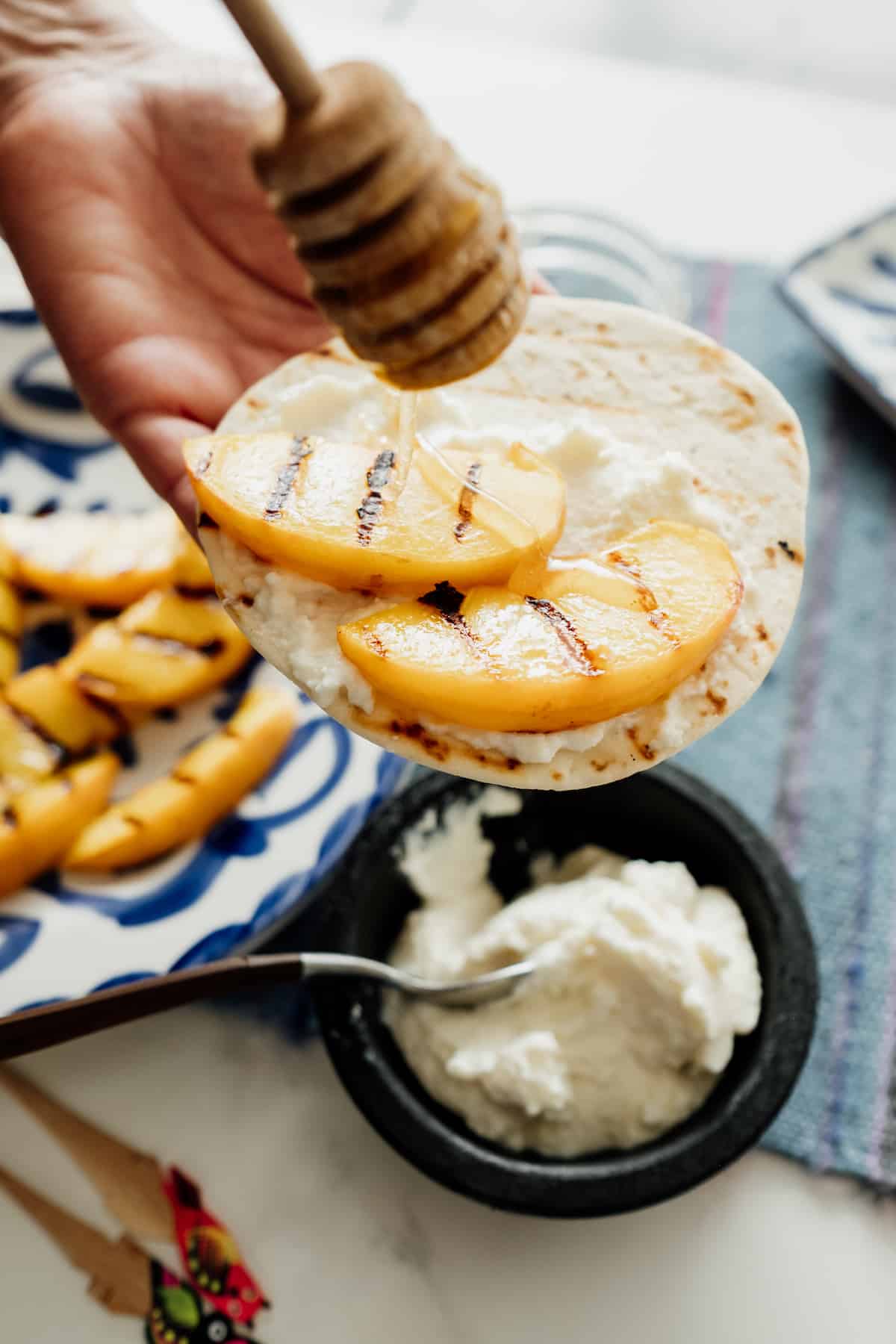 a hand holding a flour tortilla smothered with ricotta and grilled peaches drizzling honey on top. A side of plate with additional grilled peaches
