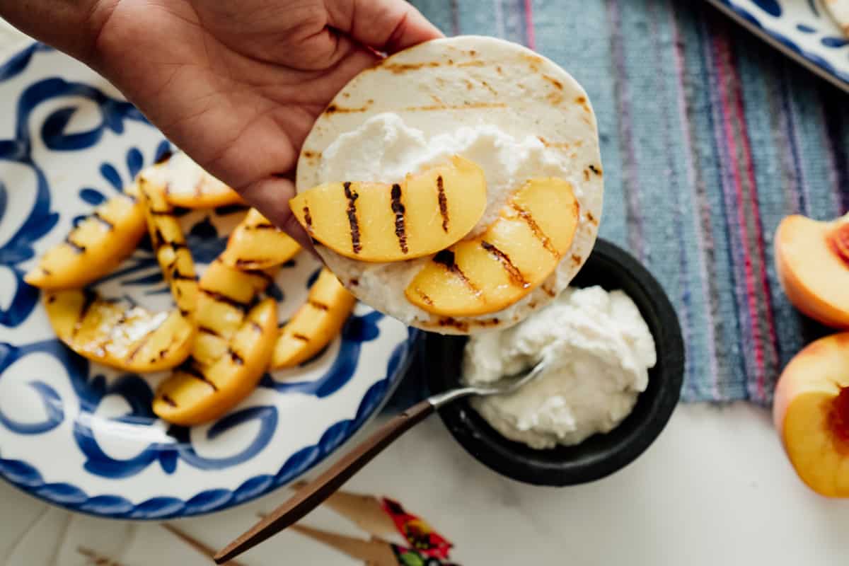 a hand holding a flour tortilla smothered with ricotta and grilled peaches. A side of plate with additional grilled peaches