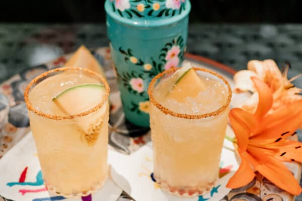 two glasses of melon margarita on a serving tray with a turquoise cocktail shaker and orange lily