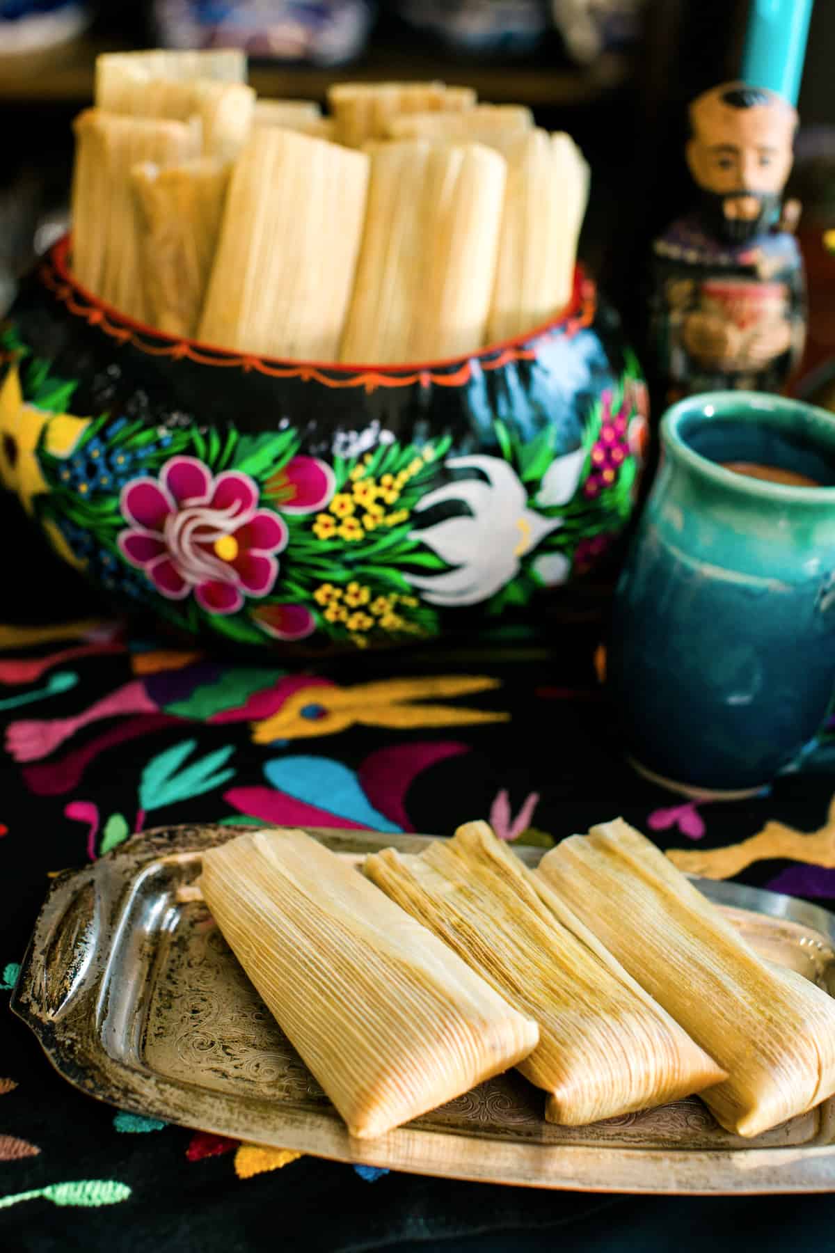 steamed pumpkin tamales on a silver serving platter with a bowl of more tamales in the background on a festive table for Dia de los Muertos