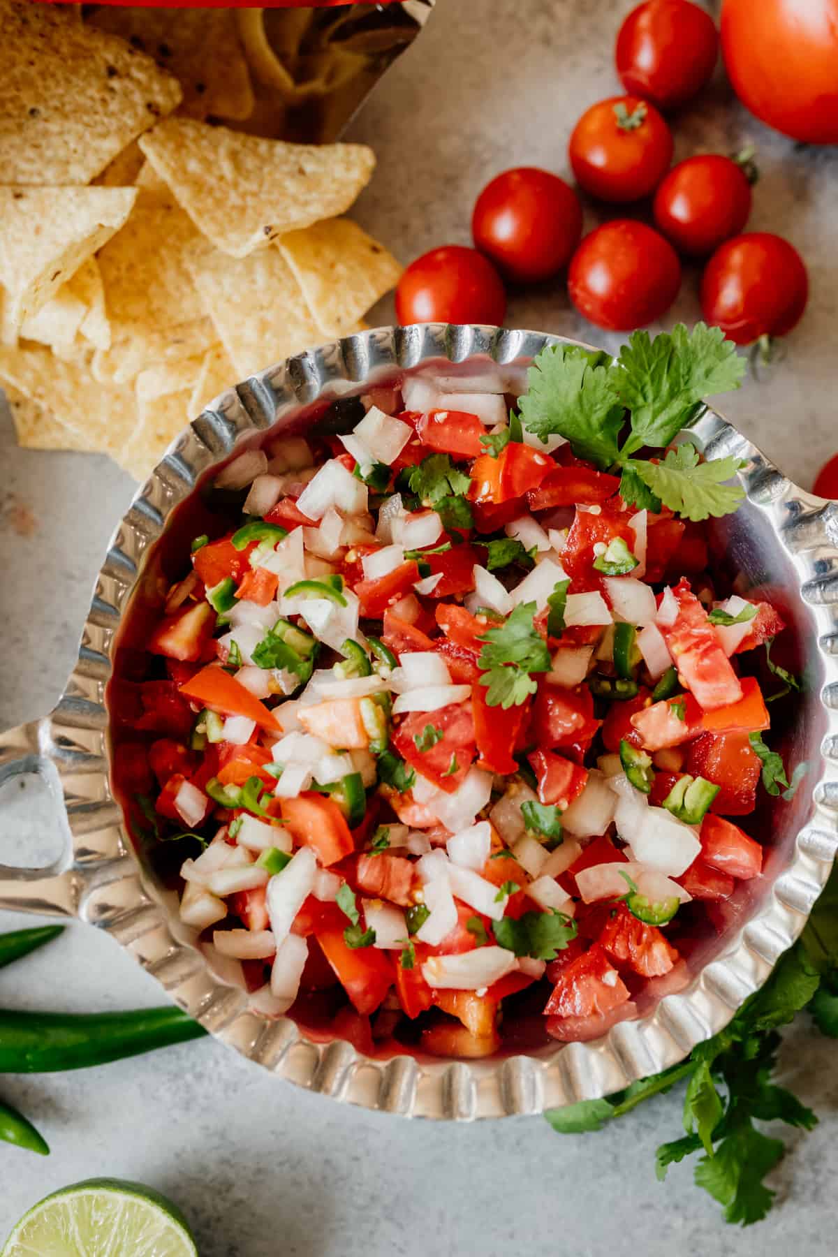 A prepared easy Pico de Gallo recipe served in a large silver bowl with tortilla chips and remaining chopped tomato salad ingredients placed around it