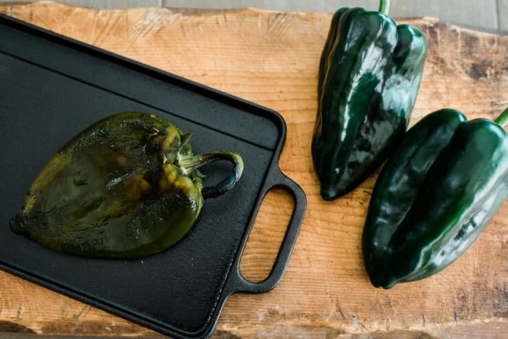 roasted poblano pepper on a cast iron griddle next to 2 fresh chiles poblanos. 