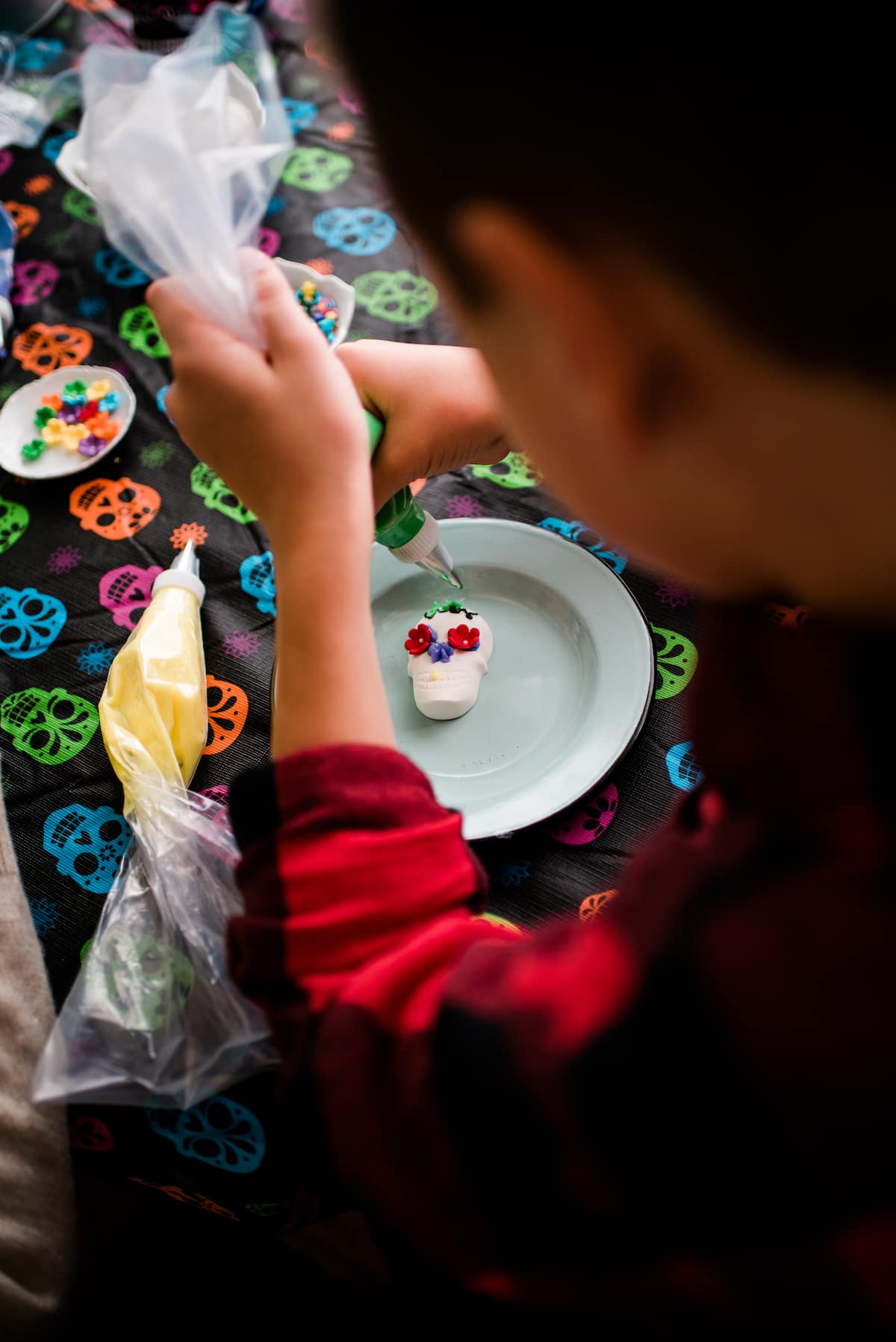 An overhead shot of a young boy decorating his Dia de los Muertos candy skulls with colorful frosting, candy flowers, and sprinkles on top of a colorful skull tablecloth 