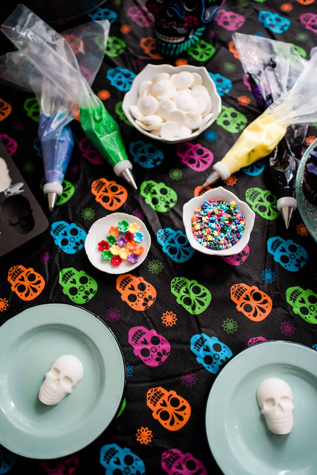 Undecorated white chocolate skulls on blue plates with piping bas filled with colorful frosting, candy flowers, and sprinkles on top of a colorful skull tablecloth 