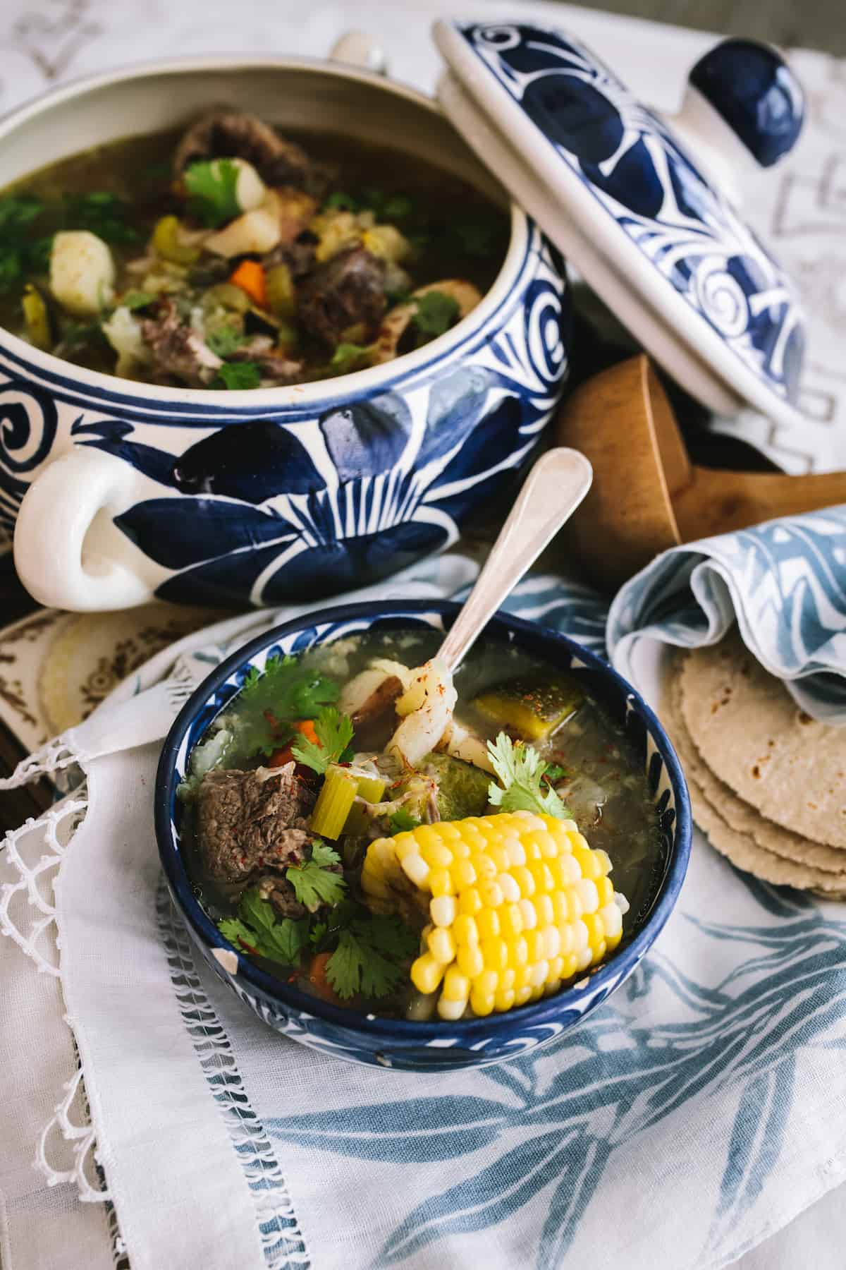 blue and white Mexican soup tureen filled with oxtail soup and a stack of corn tortillas and a wooden ladle 