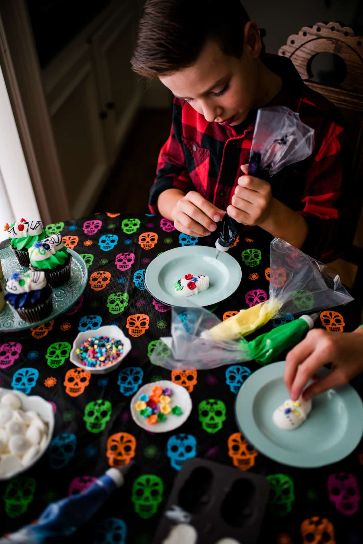The author's children decorating their Dia de los Muertos candy skulls with colorful frosting, candy flowers, and sprinkles on top of a colorful skull tablecloth 