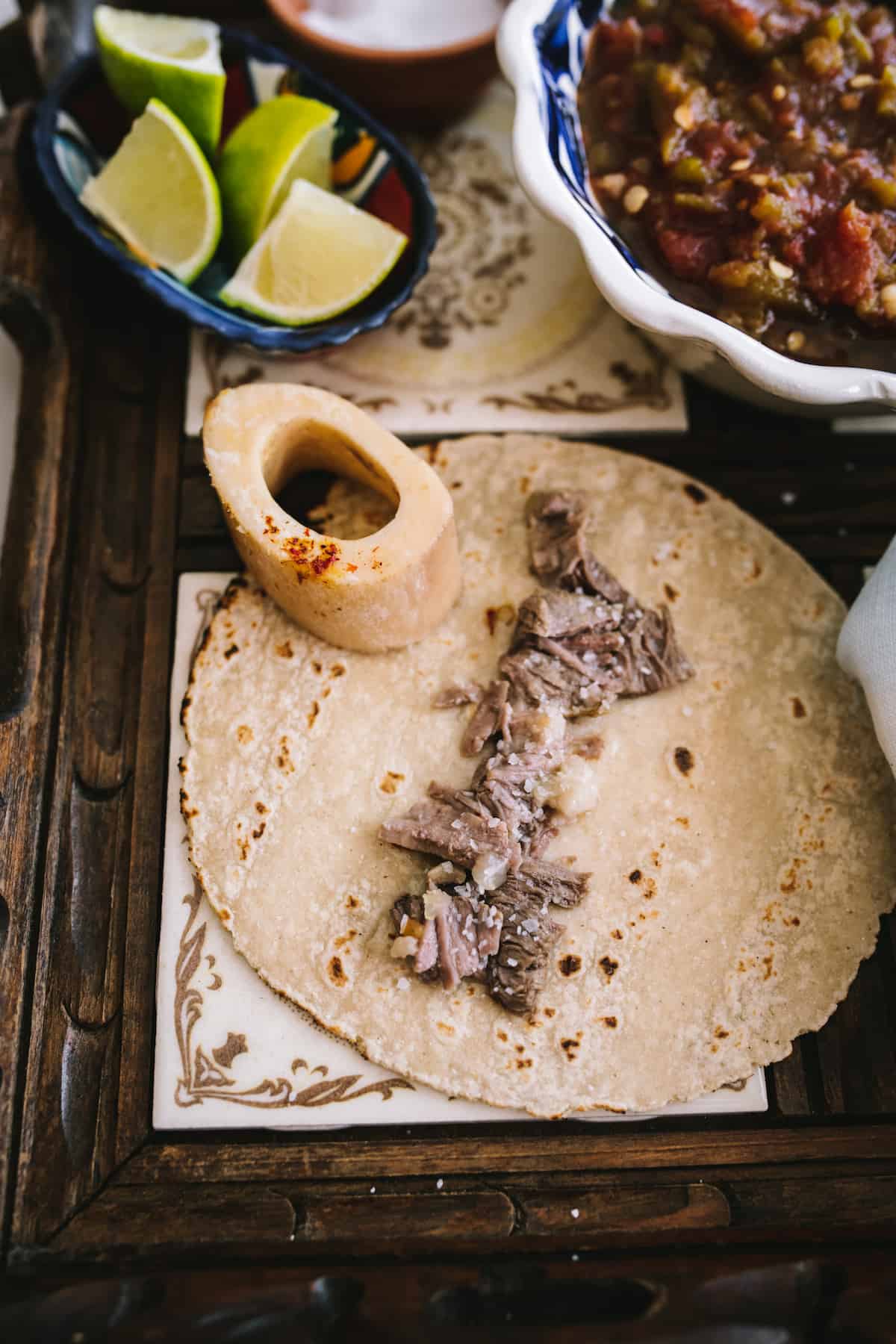 slow cooked beef and bone marrow from oxtail soup served in a corn tortilla sprinkled with salt with lime wedges in a bowl in the background