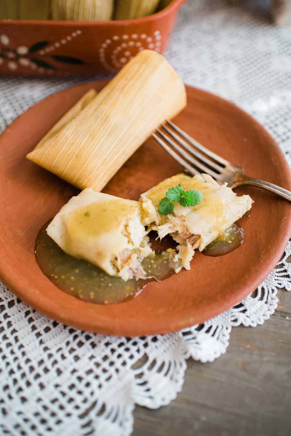 round terracotta plate with an unwrapped chicken tamale broken in half and drizzled with salsa verde with another tamale still in the husk.