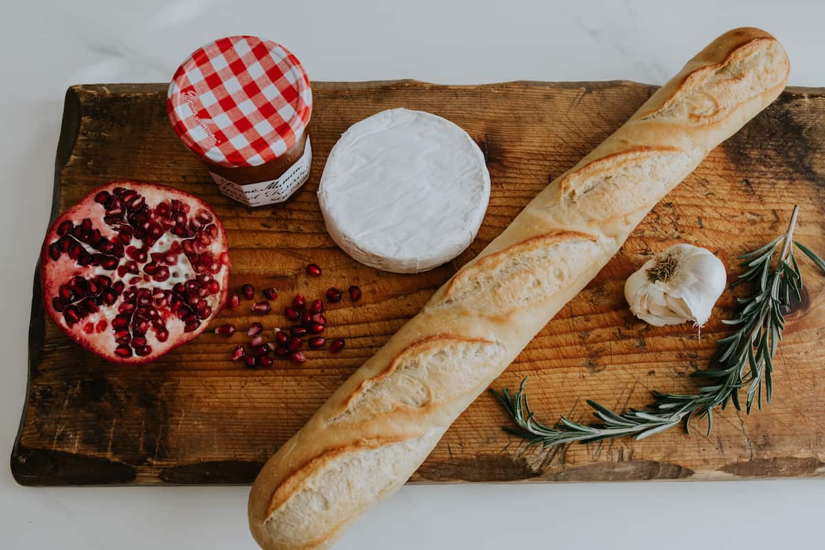 rectangular wooden board topped with a jar of bonne maman apricot preserves, a halved pomegranate, a round of brie, a baguette, a head of garlic and a sprig of fresh rosemary.