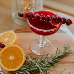 A festive cranberry margarita served with a skewer of sugared cranberries and a sugared rim in a martini class on a wooden cutting board with rosemary and orange slices as garnish. The perfect Christmas cocktail or Thanksgiving drink!