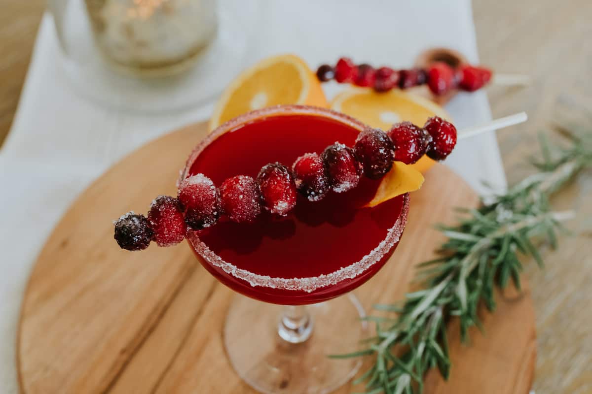 A festive cranberry margarita served with a skewer of sugared cranberries and a sugared rim in a martini class on a wooden cutting board with rosemary and orange slices as garnish. The perfect Christmas cocktail or Thanksgiving drink!