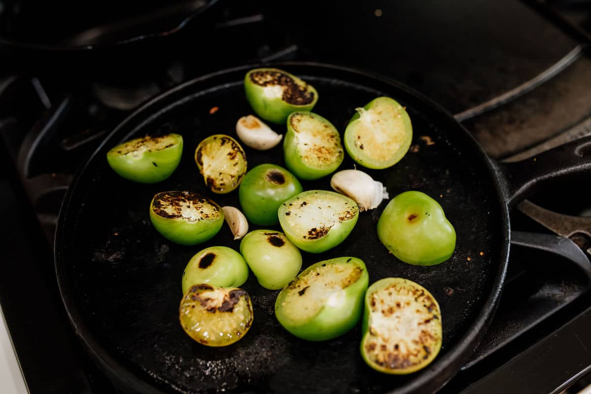 tomatillos cut in half and charred and cloves of garlic on a black cast iron skillet. 