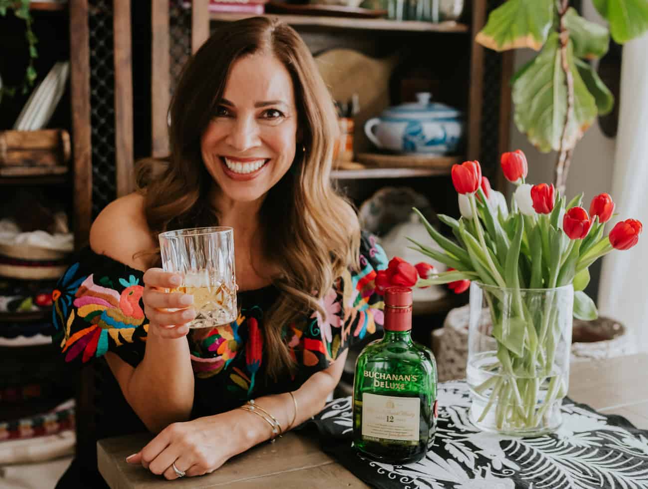 Yvette Marquez Latina Colorado food blogger holding a rocks glass of whisky with a bottle Buchanan and vase of tulips on the side.