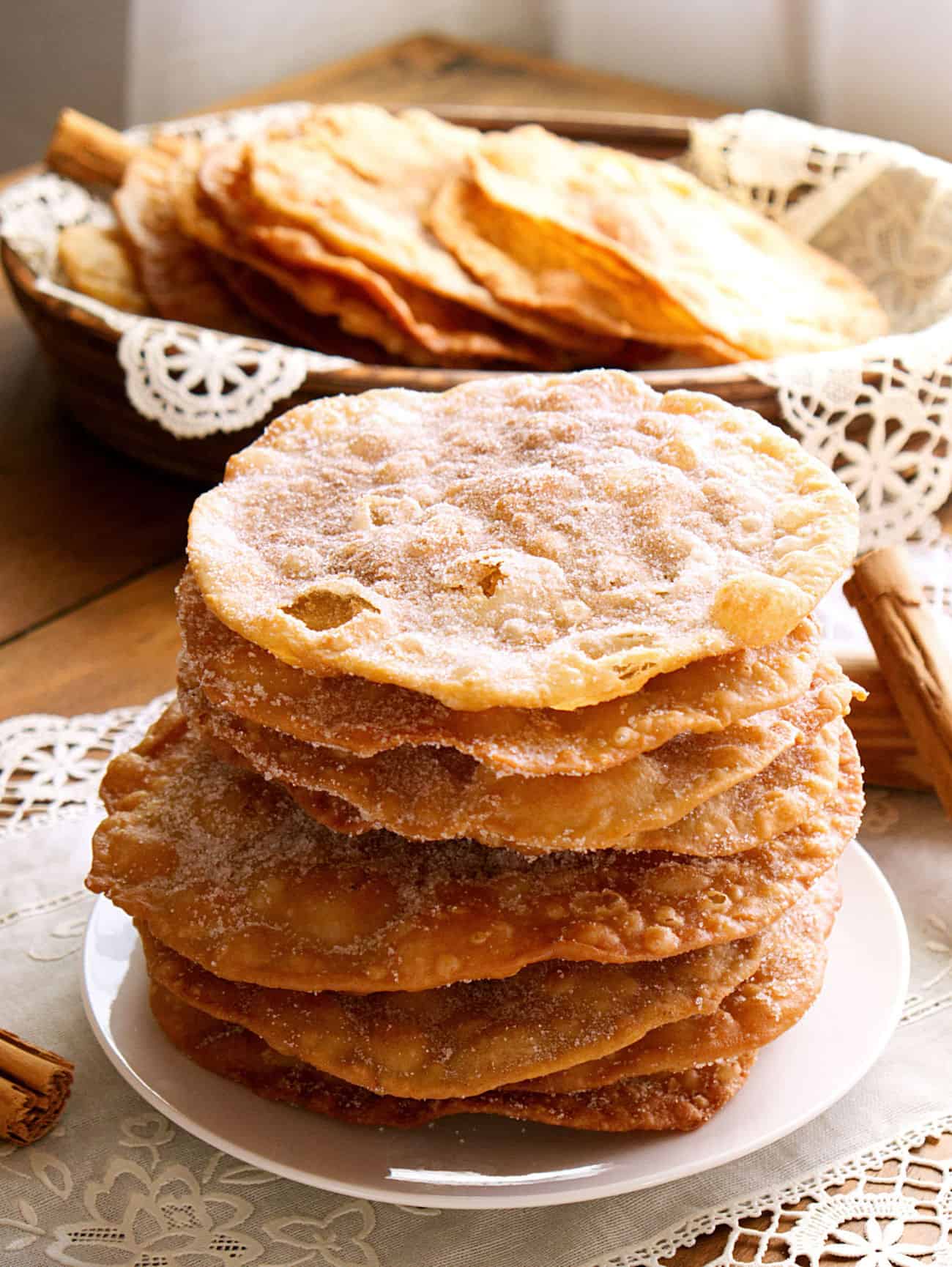 authentic bunuelos (Mexican fritters) dusted with a cinnamon sugar piled high on a plate with a basket behind with even more buñulelos.