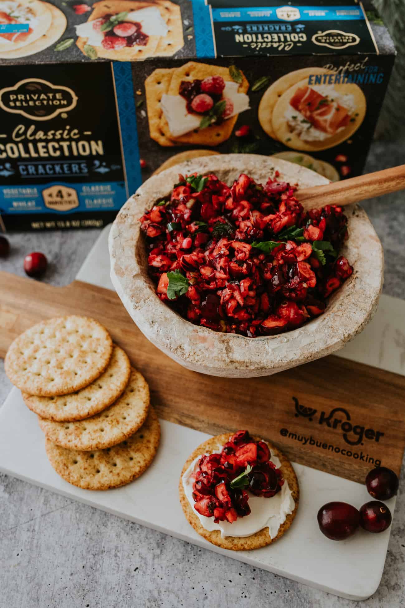 kroger branded cheese board with a bowl of cranberry jalapeño relish, a cracker smeared with cream cheese and cranberry dip, and a handful of crackers on the side.