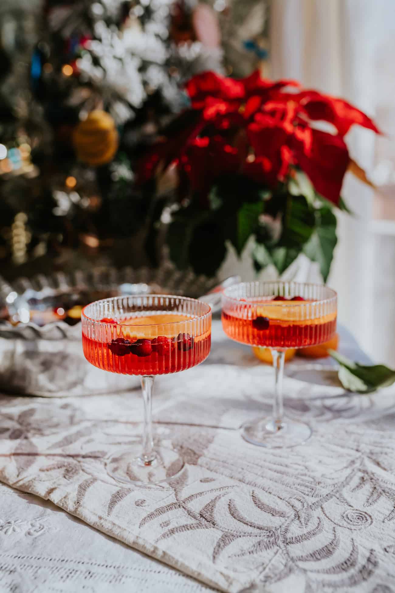 two coupe glasses filled with festive poinsettia punch for Christmas garnished with orange slices and cranberries on a white table scape with a poinsettia and a Christmas tree in the background