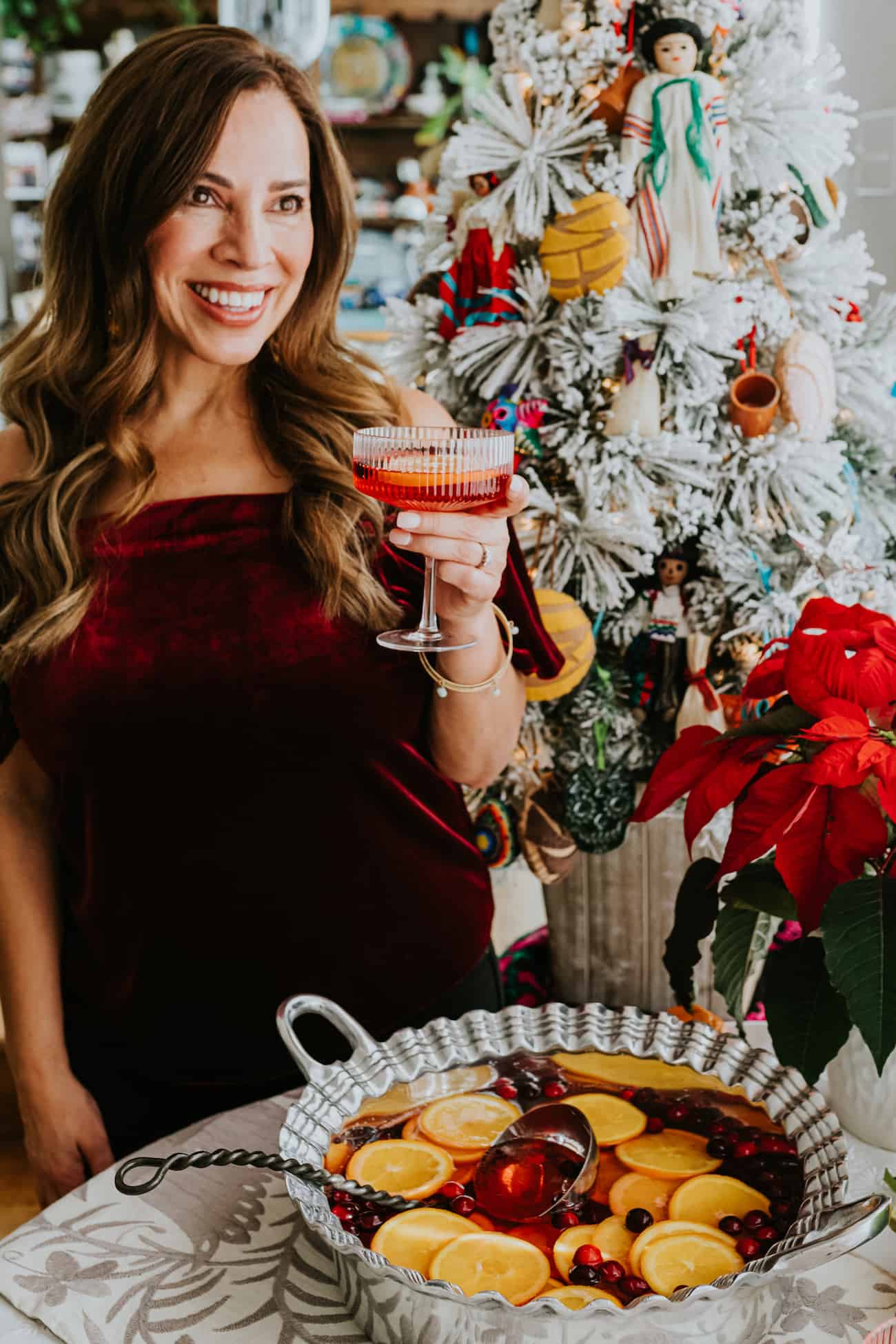 the author posing with a glass of this Christmas punch with a bowl of the holiday punch garnished with orange slices and cranberries, a potted poinsettia, and a decorated Christmas tree in the background