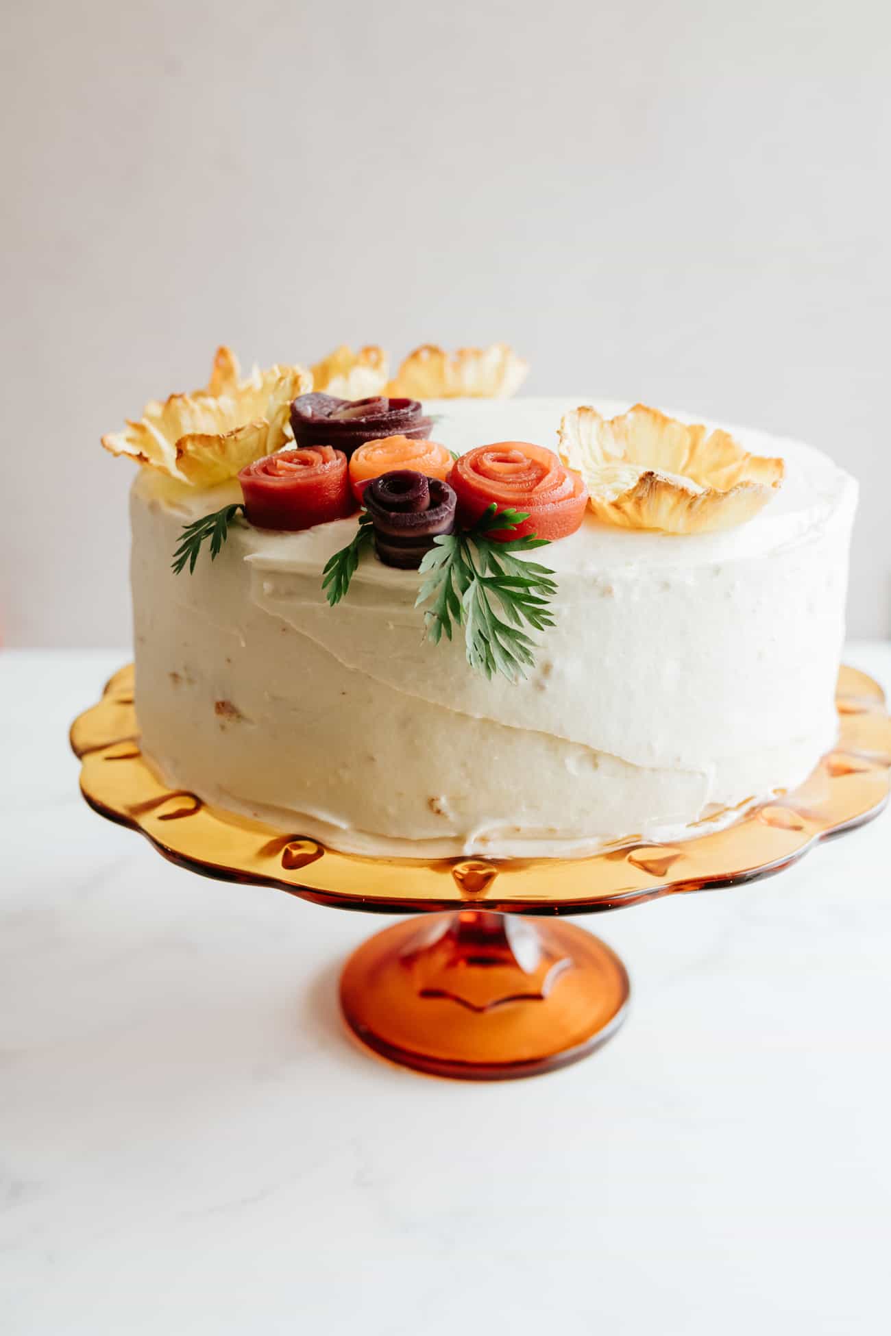 whole carrot cake decorated with pineapple flowers and rainbow carrot rosettes with carrot tops as green garnish.